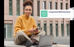 A young woman with glasses and a yellow sweater sits cross-legged outdoors with a laptop and a smartphone. An overlay on the image reads: Integrated B2B Solutions, next to a green shield icon.